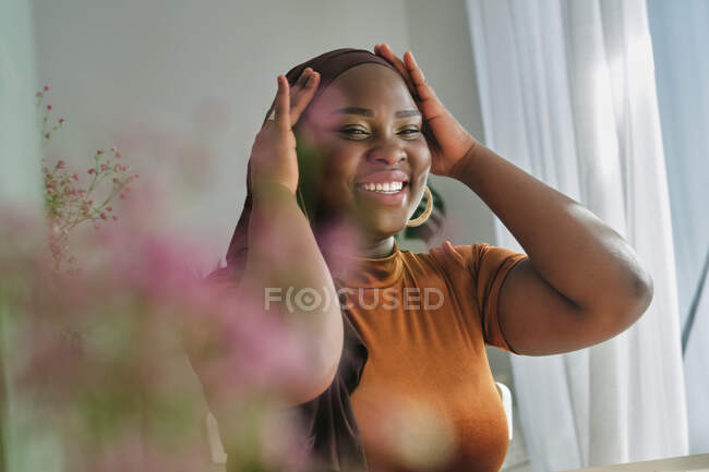 Счастливая молодая стильная африканская леди в традиционном хиджабе с руками на голове и глядя в зеркало в солнечном свете дома — стоковое фото