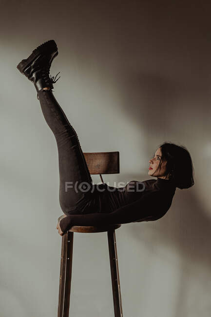 Вид збоку тонкої жінки в чорному вбранні балансує на дерев'яному табуреті з піднятими ногами в кімнаті на білій стіні — стокове фото