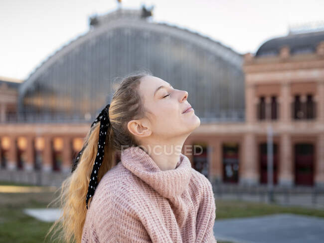Side view of daydream teenage female with long wavy hair in warm sweater with eyes closed standing on city square — Stock Photo