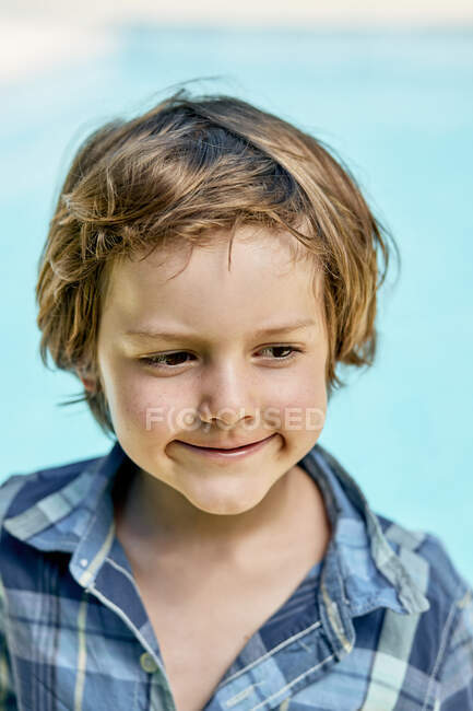 Adorable little boy with blond hair in stylish checkered shirt smiling and looking down while standing against blue background in sunlight — Stock Photo