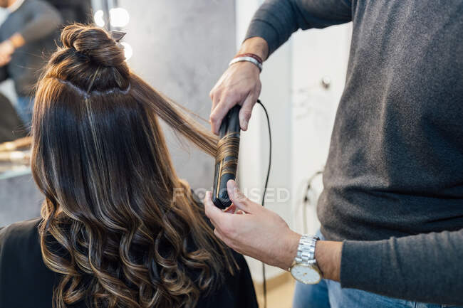 Невпізнаваний чоловічий стиліст у повсякденному одязі, використовуючи випрямлення волосся, роблячи завитки для жіночого клієнта в салоні краси — стокове фото