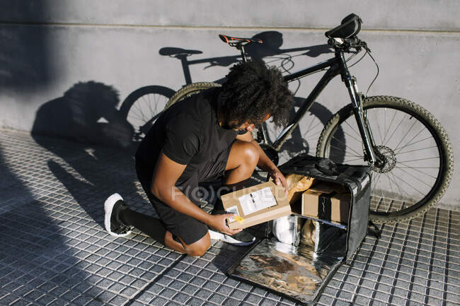 Сверху вид черного курьера с картонной коробкой, приседающего на дорожке рядом с велосипедом под солнечным светом — стоковое фото