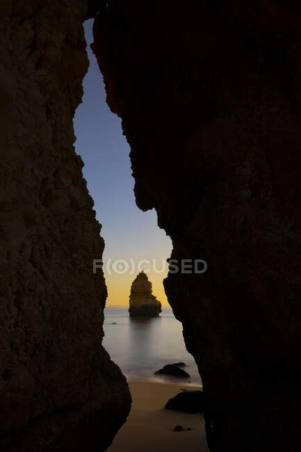 Através de caverna rochosa de pitoresca praia de areia do oceano contra o céu do pôr-do-sol na Praia do Camilo no Algarve, Portugal — Fotografia de Stock