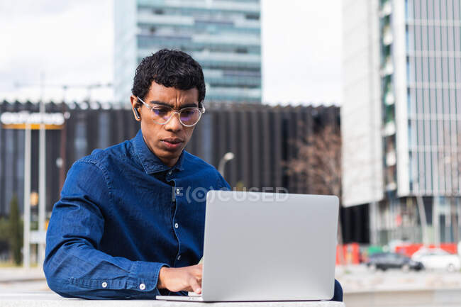 Adult concentrated ethnic male remote employee in earbud working on netbook in town on sunny day — Stock Photo