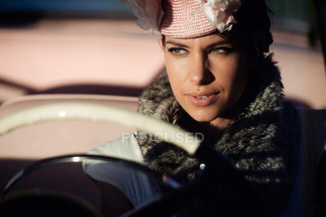 Cosecha femenina de moda con flor en el sombrero en el automóvil mientras mira hacia otro lado - foto de stock