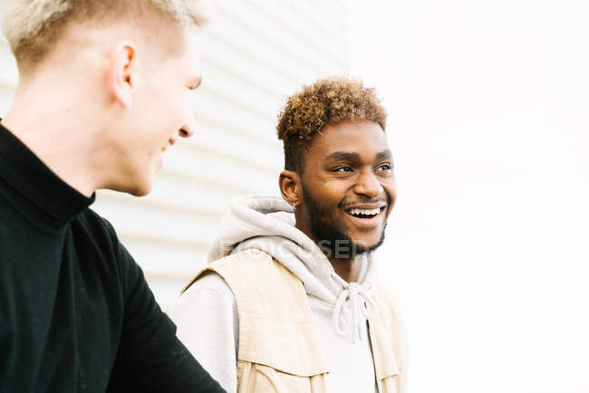 Вид сбоку на счастливых молодых друзей-мужчин разных рас в трикотажных нарядах, стоящих на городской улице возле современного здания — стоковое фото