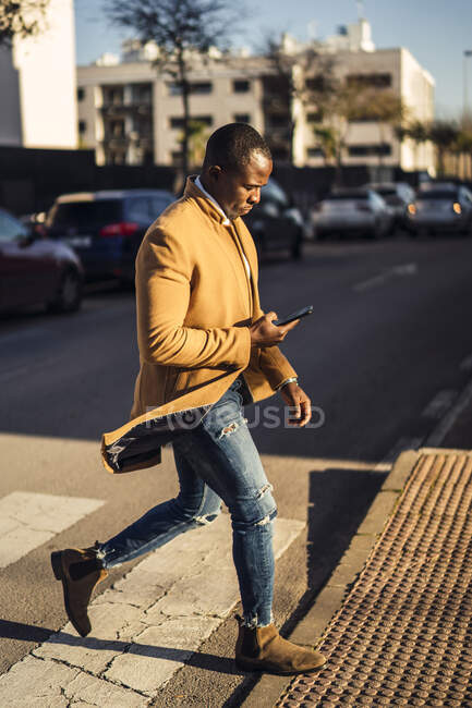 Vista laterale di grave giovane ragazzo afroamericano in abito elegante navigazione cellulare durante l'attraversamento strada nel quartiere della città — Foto stock