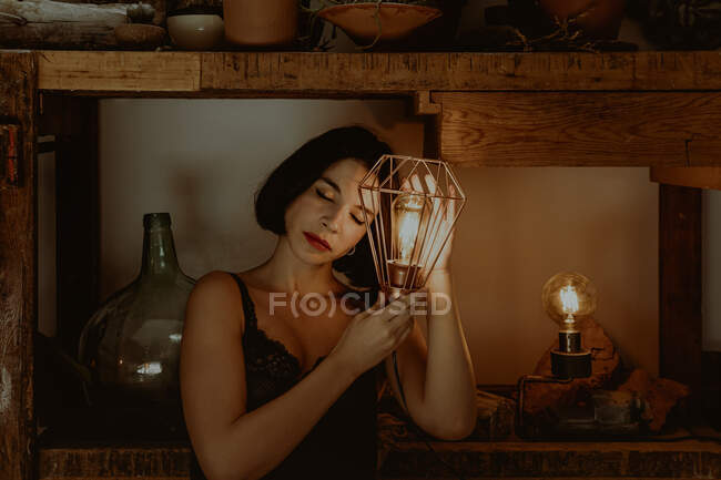 Delicate female touching illuminated metal lantern while standing in room at home with closed eyes — Stock Photo
