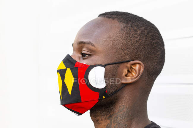 Vue latérale du jeune homme afro-américain regardant loin dans un masque lumineux avec ornement pendant la période du coronavirus sur fond clair — Photo de stock