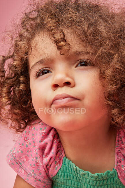 Mignon réfléchi tout-petit fille avec des cheveux bouclés dans un vêtement décontracté regardant loin sur fond rose — Photo de stock