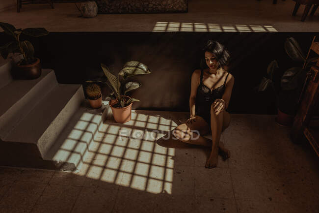 Зверху меланхолійний босоніж самка в нічній сукні сидить на підлозі в кімнаті з горщиками і сонячним світлом — стокове фото