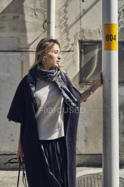 Молодая созерцательная женщина в модной одежде смотрит в сторону от столба в городе в солнечный день — стоковое фото
