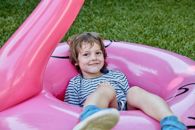 Corps complet de gai petit garçon en vêtements décontractés couché sur flamant rose gonflable tout en s'amusant sur pelouse herbeuse dans le parc — Photo de stock