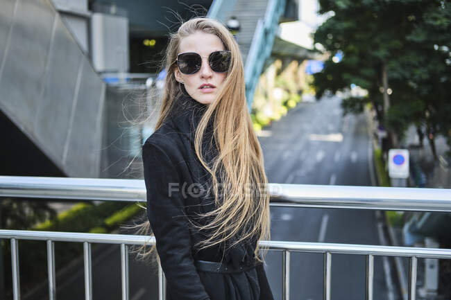 Vista lateral da jovem fêmea elegante em óculos de sol e roupa preta na ponte sobre a estrada da cidade — Fotografia de Stock
