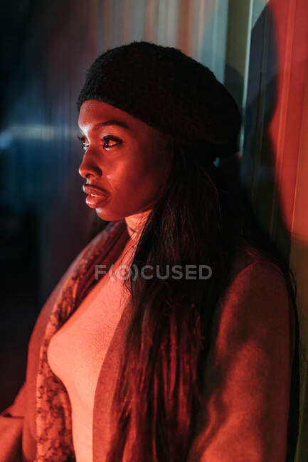 Side view of stylish African American female glowing by red neon light standing near building in city and looking away — Stock Photo