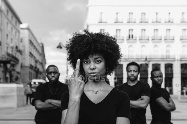 Black and white of young rude ethnic female social justice warrior demonstrating fuck gesture near self assured male partners looking at camera - foto de stock