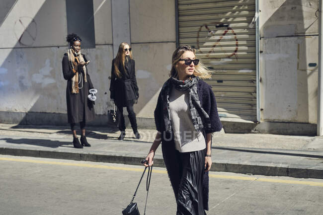 Jovem fêmea loira em roupas pretas elegantes e óculos de sol na estrada da cidade contra diversas amigas femininas nas costas iluminadas — Fotografia de Stock
