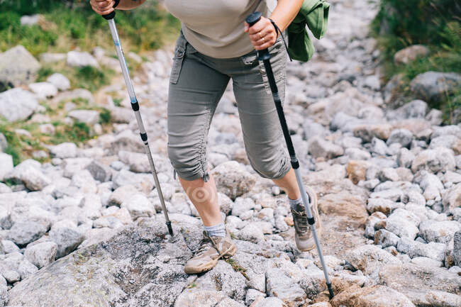 Gestutzte, bis zur Unkenntlichkeit erwachsene Frau in Freizeitkleidung übt sich im Stangenlaufen und geht auf steinigem Pfad am Berghang in sommerlicher Natur — Stockfoto