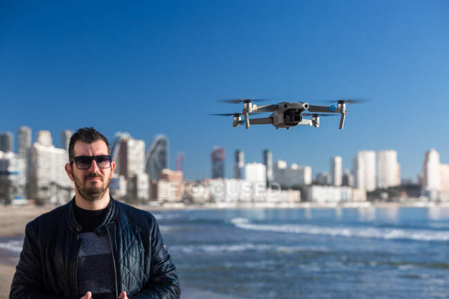 Schwerer Mann mit Sonnenbrille und Fernbedienung steht auf Drohne am Strand in der Nähe der Stadt — Stockfoto