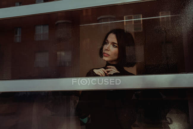 Through glass of thoughtful female standing near window and observing street while looking away — Stock Photo