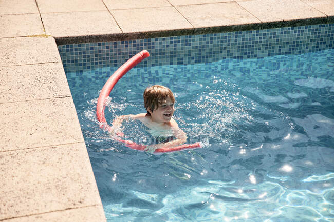 Ângulo alto do menino bonito atencioso que jaz na placa do pontapé do treinamento ao nadar na piscina exterior no dia ensolarado — Fotografia de Stock