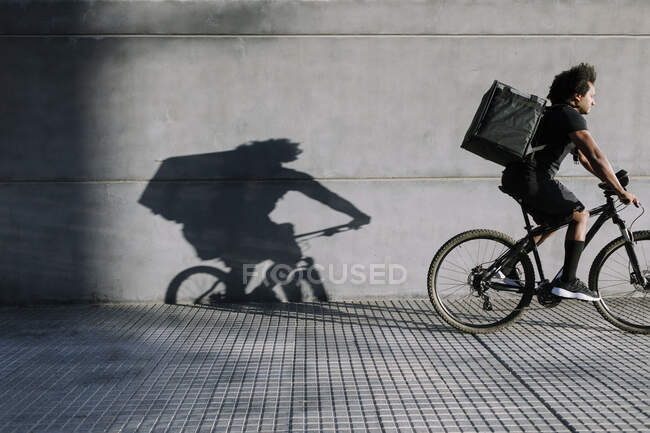 Side view of ethnic male with delivery box riding bicycle on walkway against wall with shadow in sunlight — Stock Photo