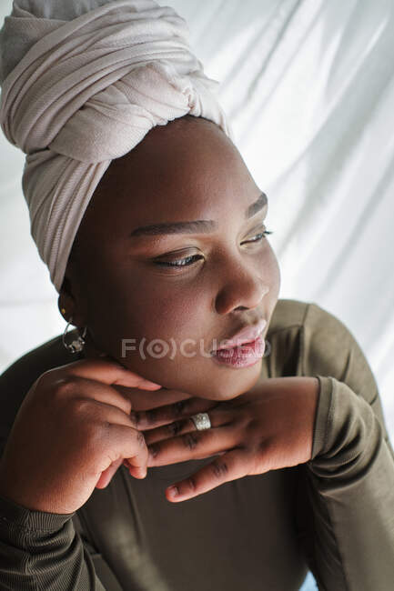 D'en haut de séduisante jeune femme noire en vêtements décontractés et turban assis sur la chaise avec les mains au menton et regardant loin — Photo de stock