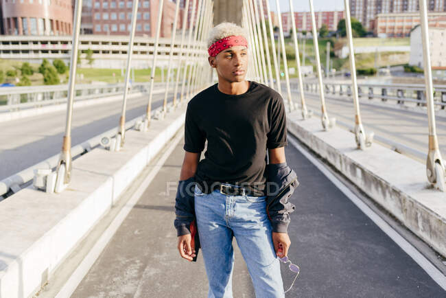 Guapo chico negro con pañuelo en la cabeza de pie en el medio del puente mirando hacia otro lado - foto de stock