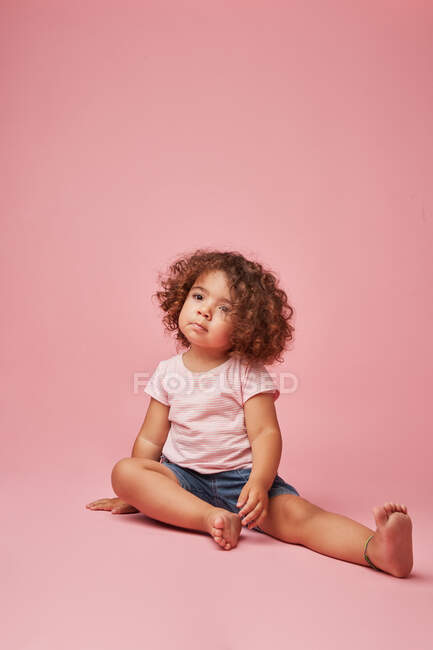 Милая вдумчивая девочка с вьющимися волосами в повседневной одежде смотрит в сторону, сидя на розовом фоне — стоковое фото