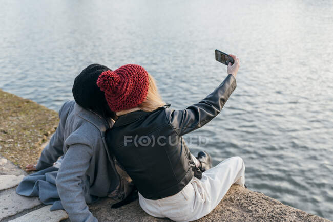 Mujer irreconocible tomando selfie de amiga negra mientras se relaja en terraplén en la ciudad - foto de stock
