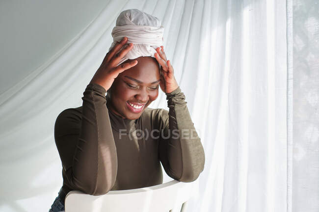 Вид сбоку на привлекательную молодую черную леди в повседневной одежде и тюрбан, сидящую на стуле с руками на голове и закрытыми глазами — стоковое фото