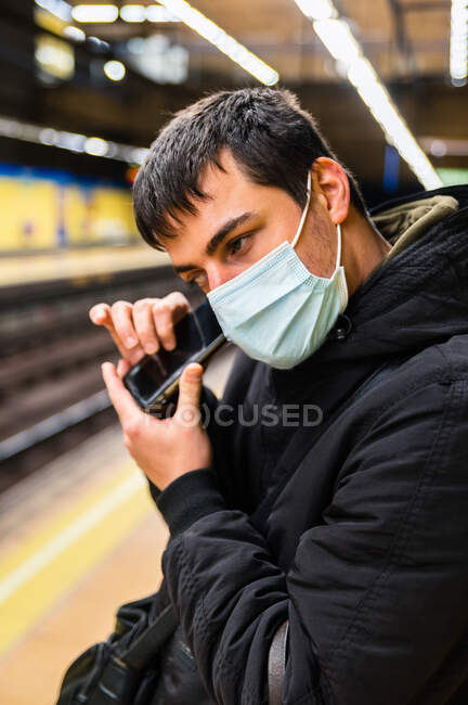 Vista laterale del passeggero cieco in maschera medica in attesa di treno alla piattaforma e altoparlante di ascolto sul telefono cellulare — Foto stock