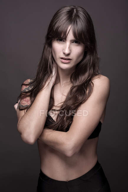 Sensual female with tattoo wearing bra touching neck standing in studio against black background — Stock Photo