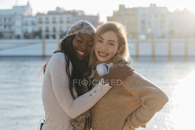 Guardando la fotocamera due amici femminili multirazziali in cuffia godendo insieme su argine nella giornata di sole — Foto stock