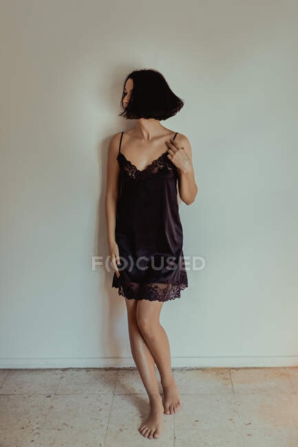 Graceful tender female in nightgown standing in room leaning against a white wall and looking away — Stock Photo