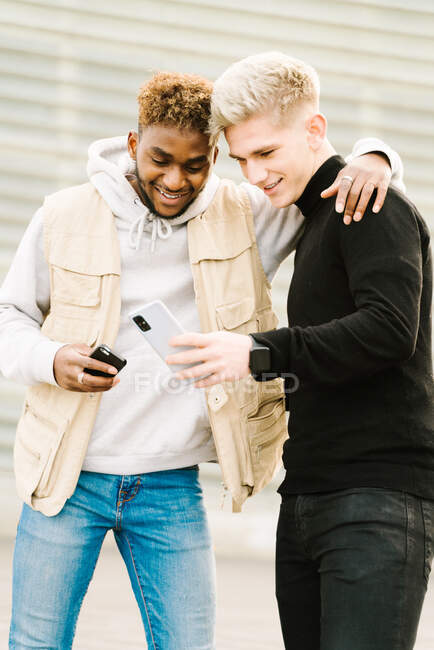 Щасливий афроамериканець у модному одязі посміхається, стоячи на вулиці з рукою на плечі друга-чоловіка, який показує відео на смартфоні. — стокове фото