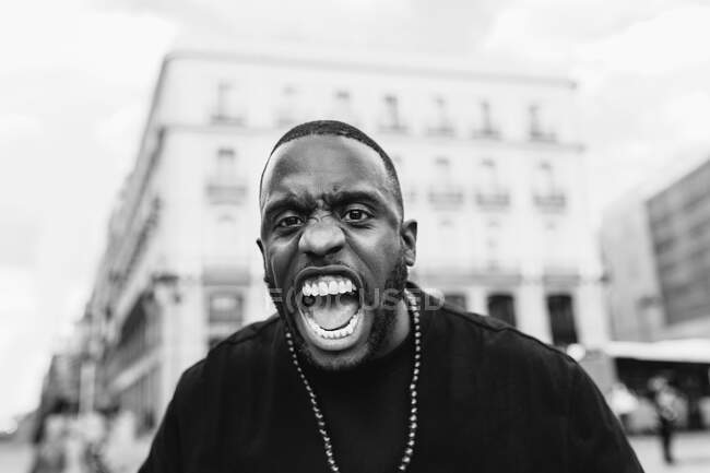 Black and white of young angry ethnic male screaming while looking at camera in town — Stock Photo