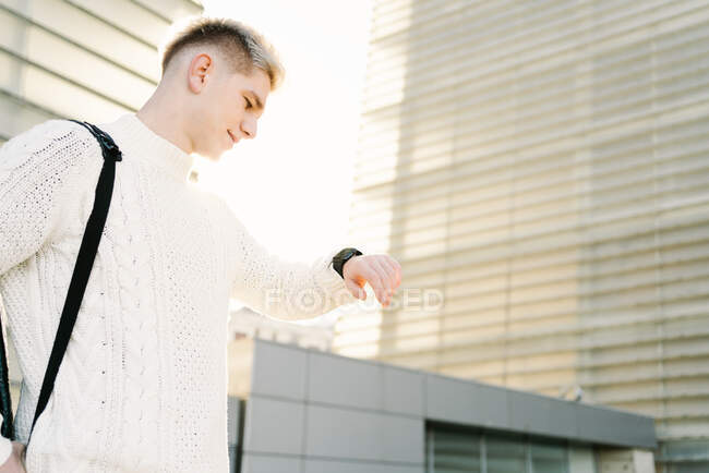 Вид сбоку молодого стильного парня в трикотажном вязаном свитере, проверяющего время на наручных часах в ожидании приема на городской улице возле современных зданий в солнечный день — стоковое фото