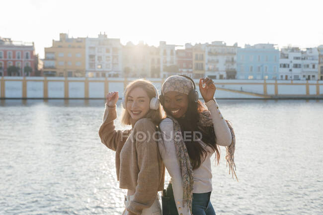 Vista laterale di amiche multirazziali in cuffia godendo della musica e ballando insieme sul terrapieno nella giornata di sole — Foto stock