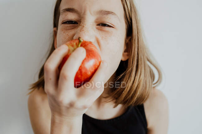 Girl in casual top looking at camera while biting fresh ripe red apple against white background — Stock Photo