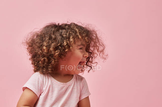 Nettes fröhliches Kleinkind Mädchen mit lockigem Haar in lässiger Kleidung schüttelt den Kopf mit geschlossenen Augen auf rosa Hintergrund — Stockfoto