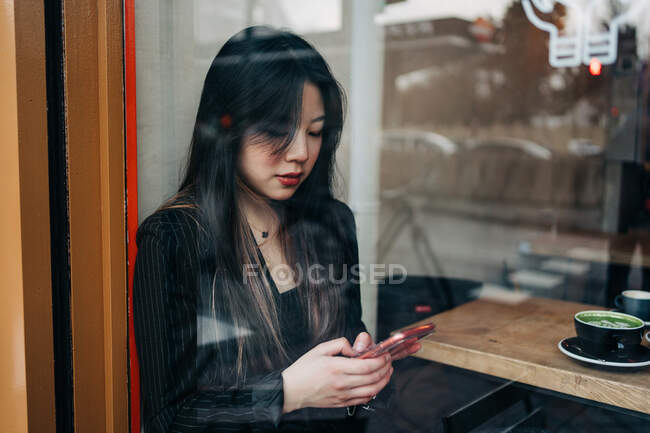 Langhaarige brünette asiatische Frau, die einen Kaffee in einem Coffeeshop trinkt, während sie ein Handy sucht — Stockfoto