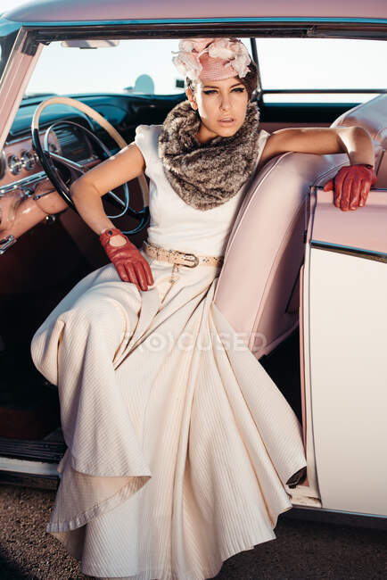 Femme féminine en chapeau avec des fleurs et des vêtements rétro avec ceinture regardant loin tout en étant assis dans l'automobile vintage — Photo de stock