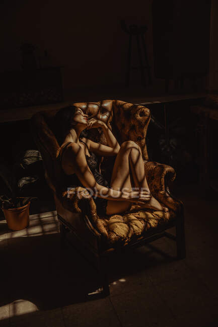 Vista lateral de mujer tranquila en camisón sentado en sillón de cuero iluminado por la luz del sol y relajante con los ojos cerrados - foto de stock