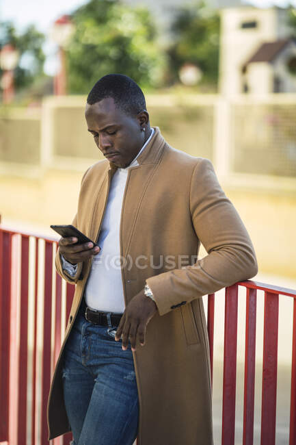 Concentrato giovane afroamericano maschio in abito alla moda appoggiato sulla recinzione sulla strada e la navigazione del telefono cellulare nella soleggiata giornata estiva — Foto stock