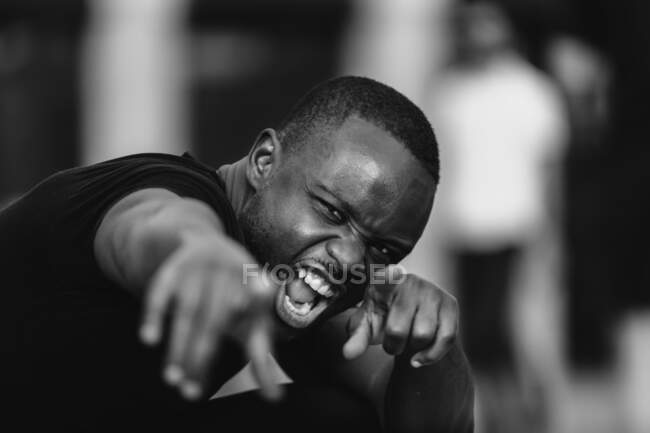 Blanco y negro de joven activista masculino étnico molesto gritando y mostrando con el dedo mientras mira la cámara en la ciudad - foto de stock