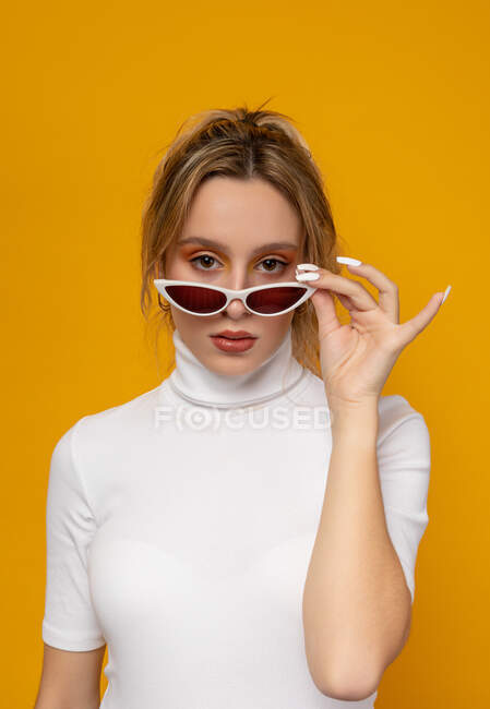 Magnifique jeune femme en vêtements blancs abaissant les lunettes de soleil à la mode et regardant la caméra tout en se tenant debout sur fond jaune en studio — Photo de stock