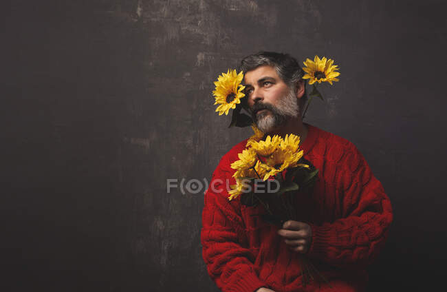 Creativo macho maduro en suéter de punto que cubre la cara con girasoles brillantes sobre fondo negro - foto de stock