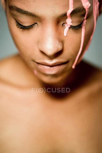 Творча етнічна жіноча модель з рожевою фарбою застрягла на її обличчі, дивлячись вниз на сірому фоні в студії — стокове фото
