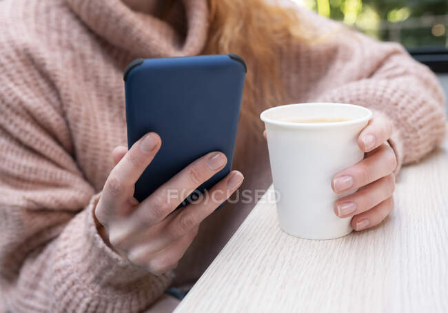 Обрізати анонімну жінку в теплому светрі, сидячи за столом з чашкою гарячого напою і використовуючи мобільний телефон — стокове фото